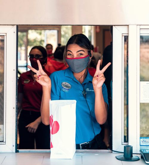 Drive-through employee wearing mask flashes a cheerful V sign gesture to customer.