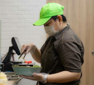 A chef in a commercial kitchen prepares a salad using tongs and a bowl.